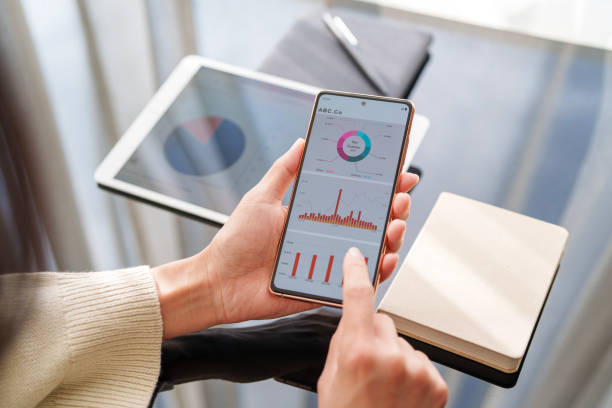 Person reviewing interactive financial dashboards on a smartphone, with tablet and notebook in background—illustrating real-time P&L reporting access.